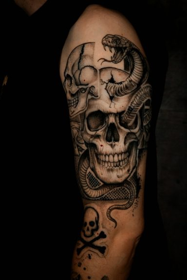 Tattoo auf dem Oberarm: Schädeltotenkopf umgeben von Schlangen und einem Piratensymbol.