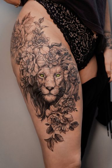 Detailreiche Tätowierung eines Löwenkopfes umgeben von Pflanzen auf dem Oberschenkel.