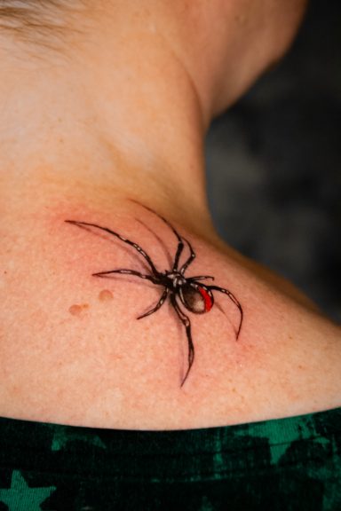 Tattoo eines schwarzen Spinnenmotivs mit rotem Punkt auf der Schulter.