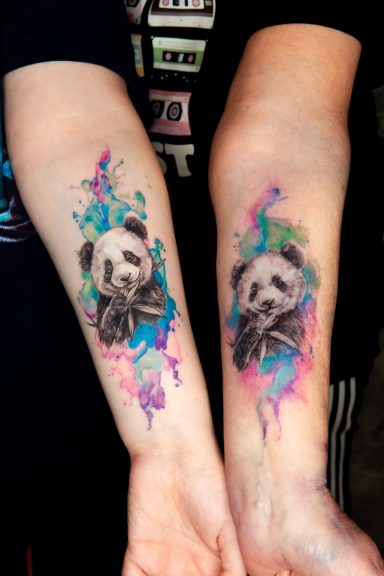 Tätowierung von einem Panda mit bunten Wasserfarben-Elementen auf beiden Armen.