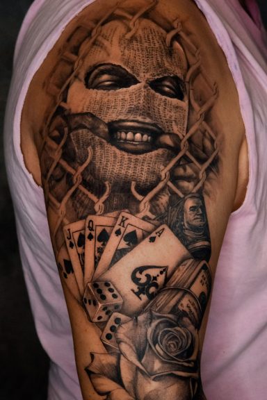 Chicano-Tattoo auf dem Oberarm mit einem Gesicht hinter einem Netz und Spielkarten.