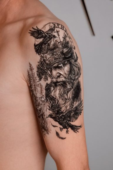 Schwarzes Tattoo eines detailreichen Männlichen Porträts und Naturmotiven auf dem Oberarm.